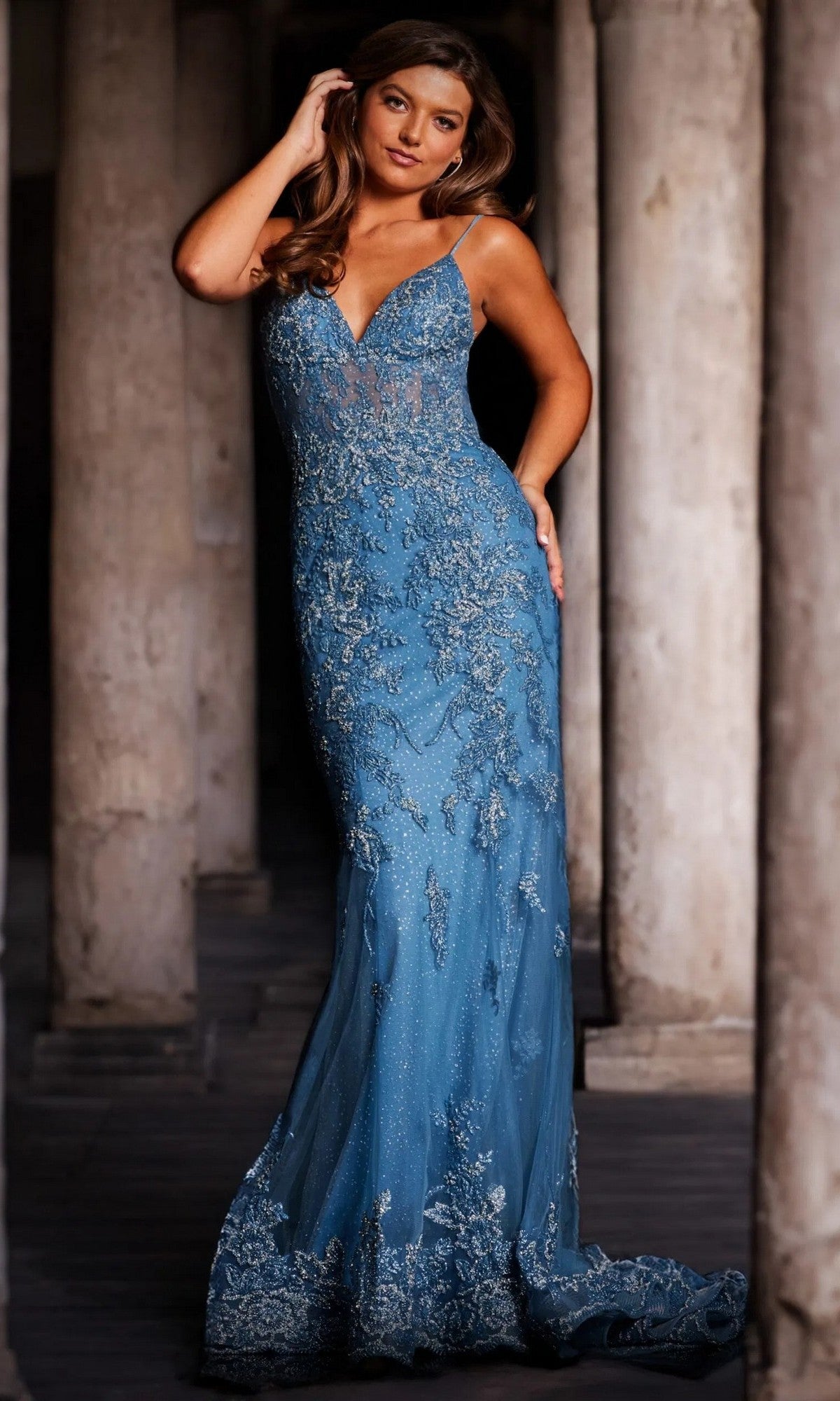A woman wearing the Jovani 37595 Sheer-Corset Long Beaded Prom Dress in blue stands by stone columns, gazing at the camera with one hand in her hair.